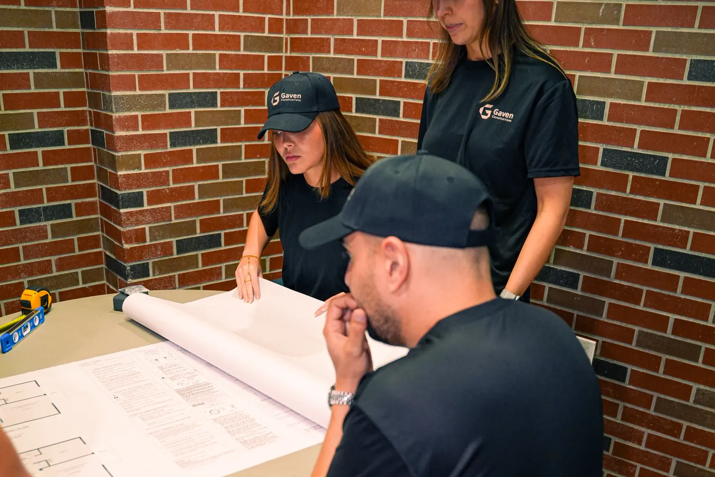 About Kitchen Remodeler Davie: the crew reading a rolled-out floor plan at the Gaven Constructions office, with a tape measure and a level on the table