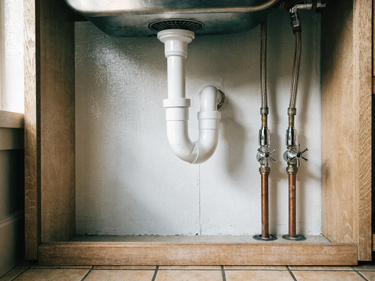 A kitchen sink base in a Davie house with both doors removed, showing the trap, the shutoff valves and the soldered copper supply lines above a dry cabinet floor