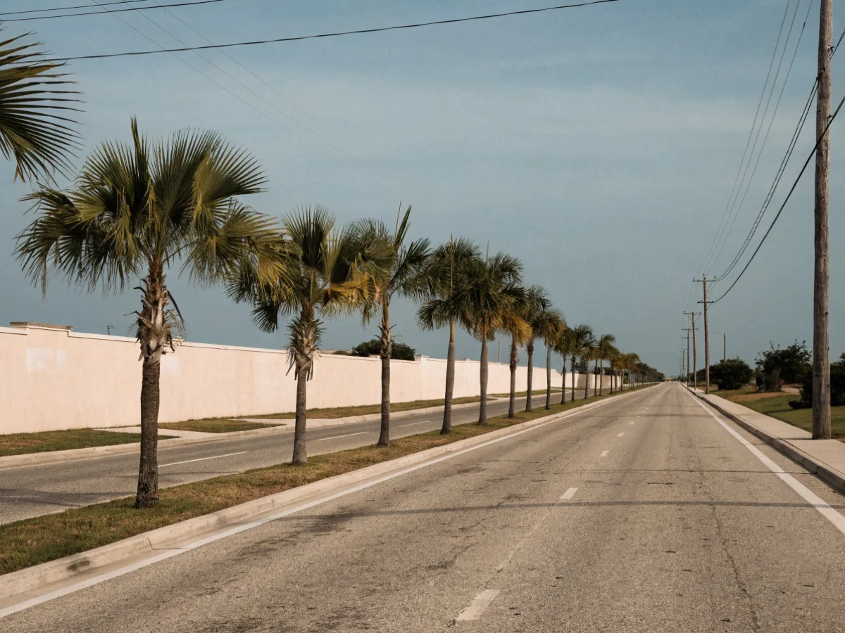 A divided residential boulevard on the west side of the school zone: two lanes each way, a planted median and a blank subdivision wall with no lettering under open sky