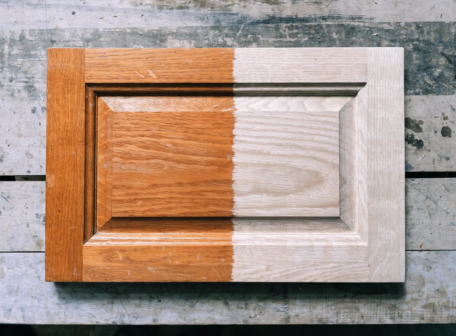 Cabinet refinishing in Davie, FL: one solid wood cabinet door photographed half stripped, old orange finish on the left half and bare sanded wood on the right, the raised panel profile running through both