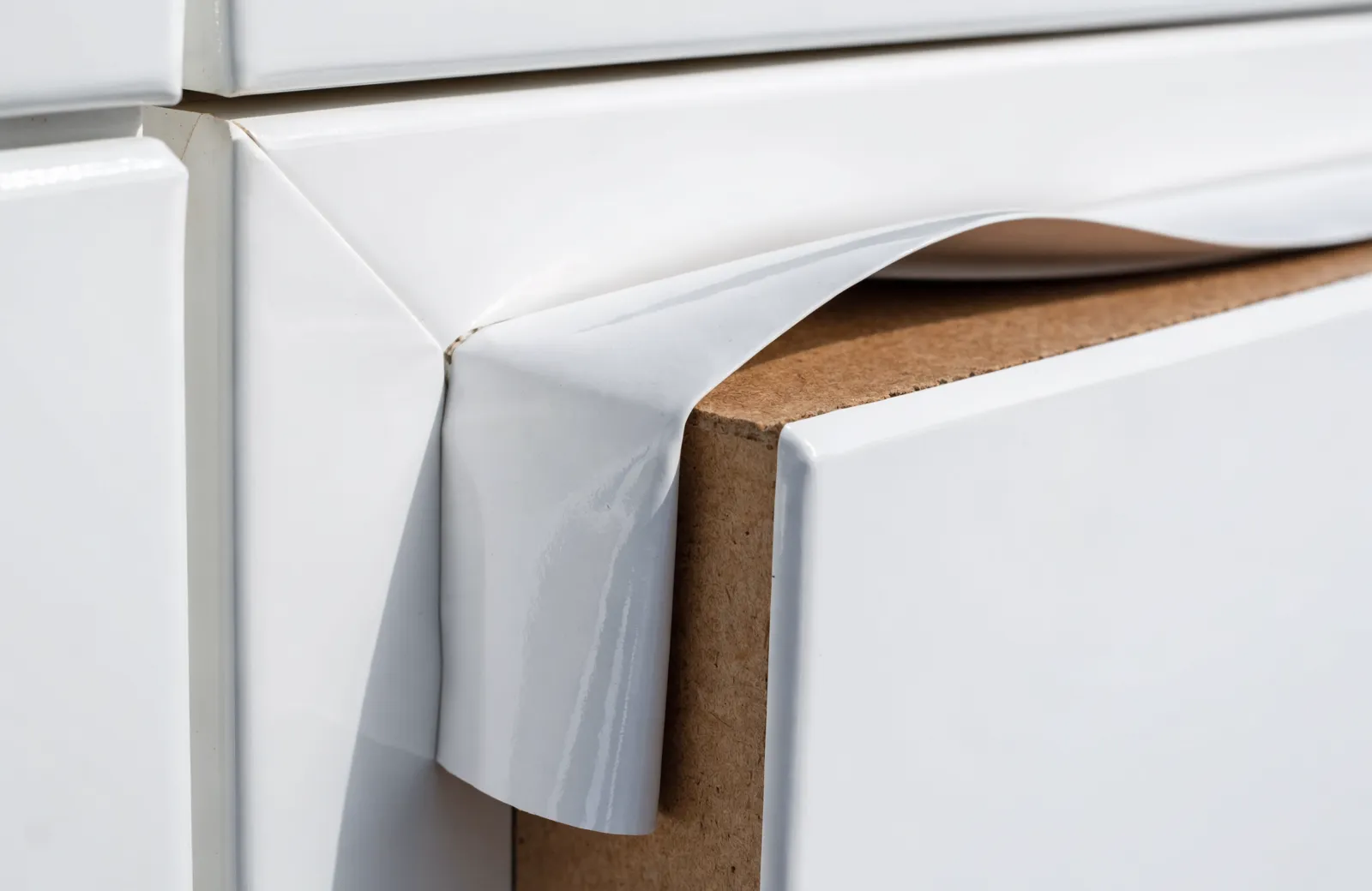 A thermofoil door with the vinyl film lifted and curled away at the top corner, exposing an MDF core that is visibly not the same material as the face