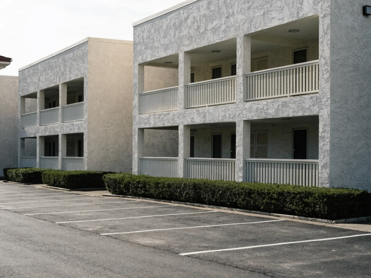 A shared parking apron in front of two story condominium blocks in Davie, painted unnumbered stalls, a clipped hedge and no individual driveway