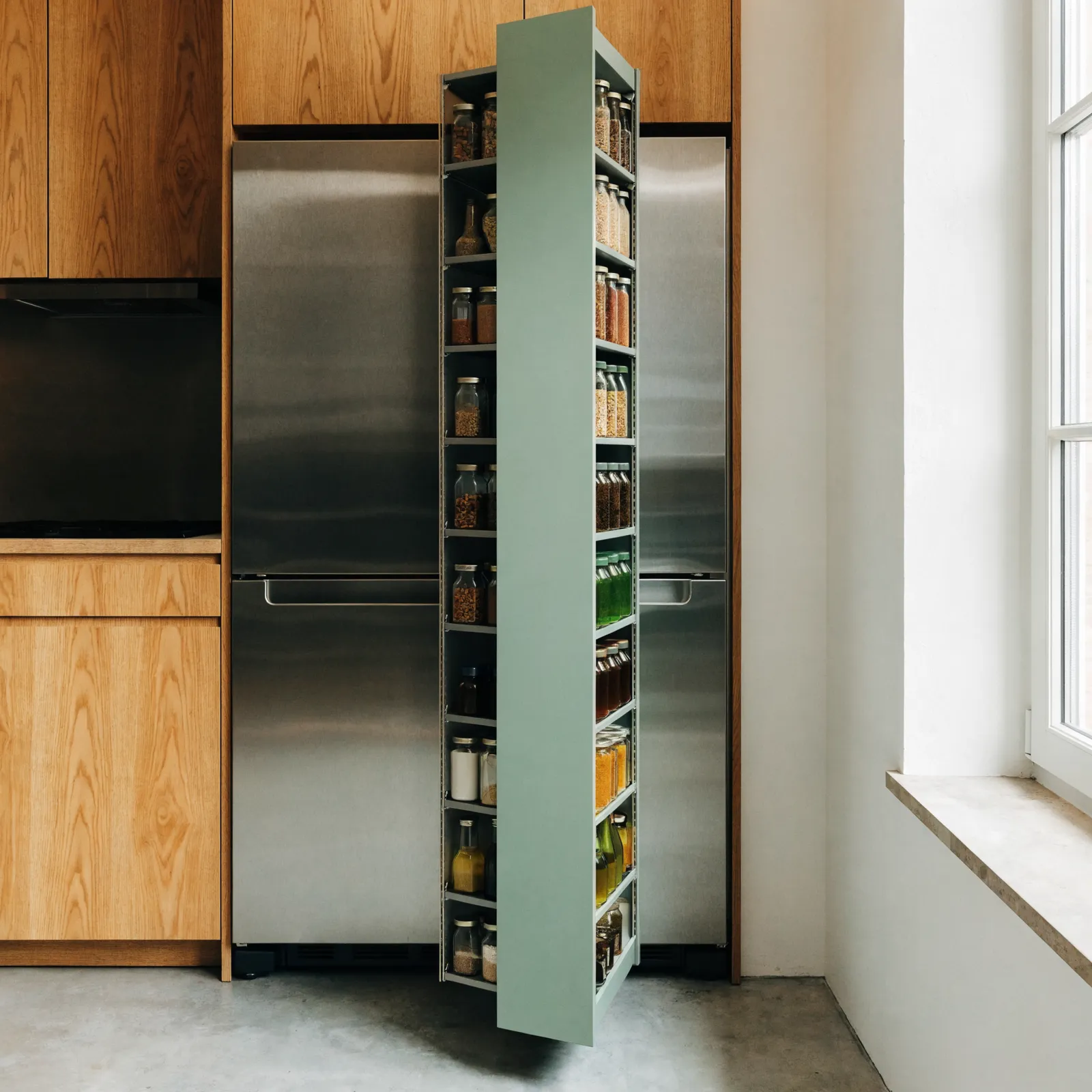 A narrow pull-out pantry column drawn out of the gap beside a refrigerator, tall and shallow, with jars and bottles on both faces
