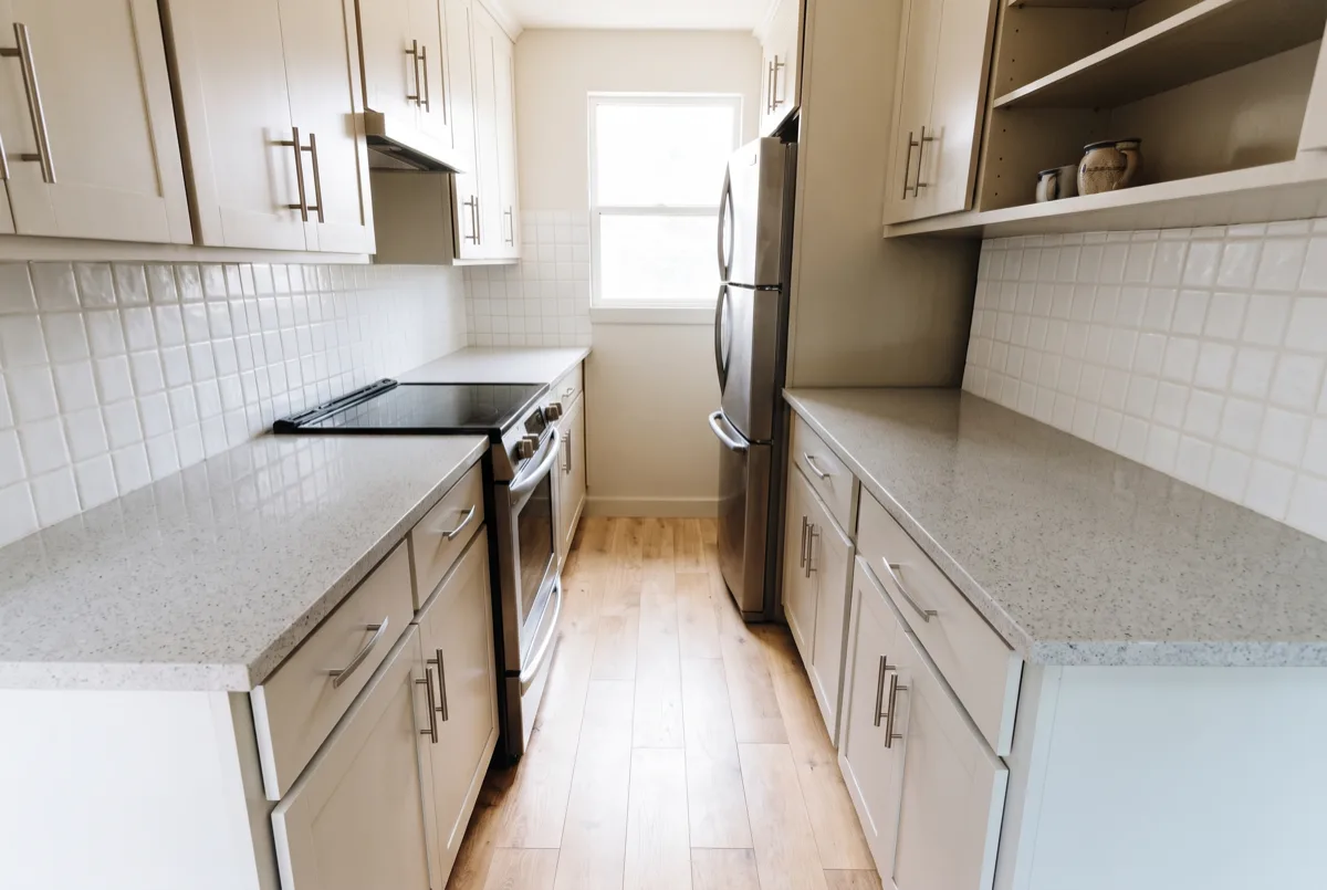 The aisle of that Davie kitchen seen straight down its length, both counter edges running away toward the window at the far end and the floor planks laid the same direction between them