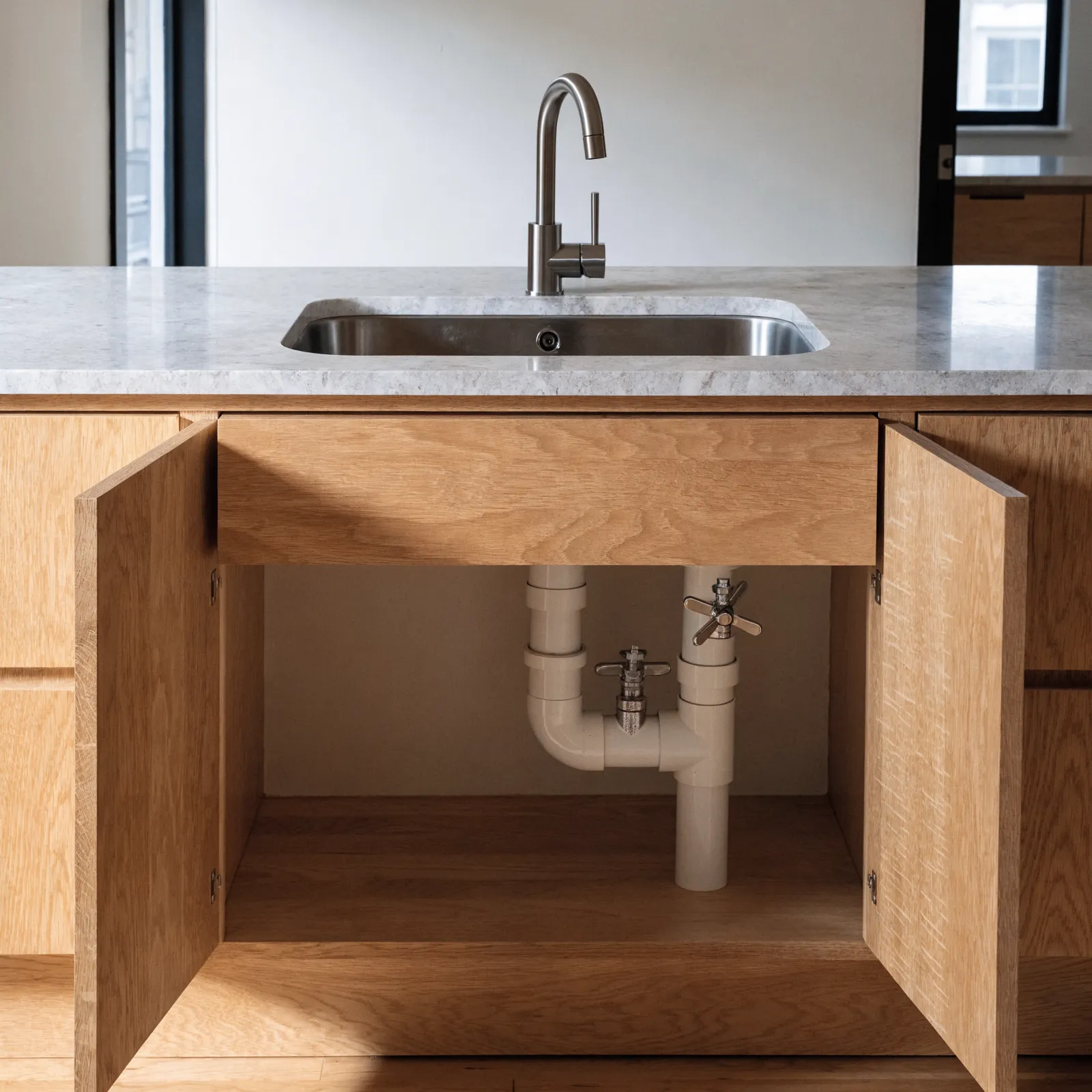 A working island: a sink base in the middle of the run with its doors open, the trap and the shutoff valves reachable behind the false drawer front