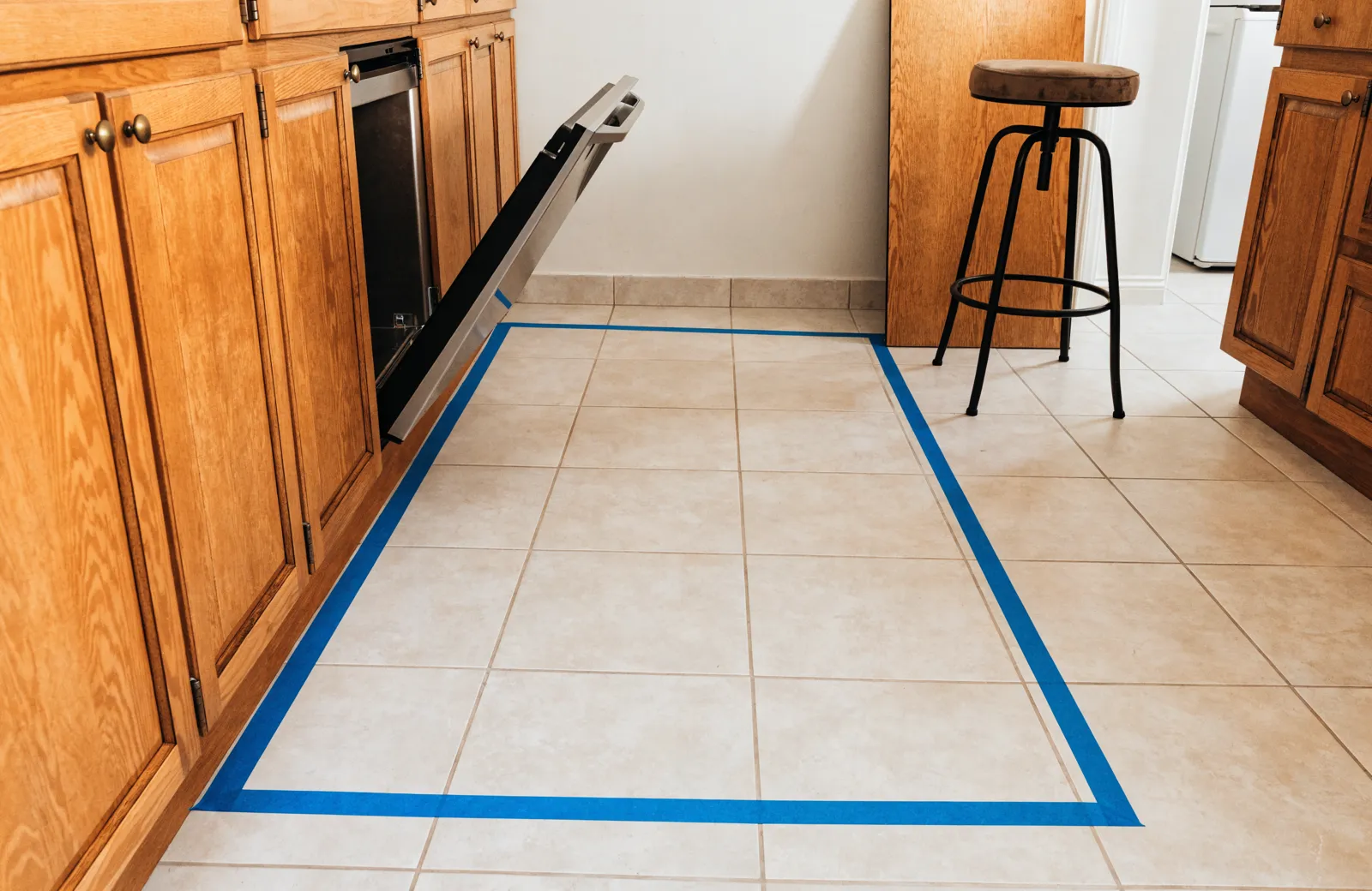 The outline of a proposed island taped onto a kitchen floor, with the dishwasher door standing open into the taped aisle and a stool pulled back on the other side