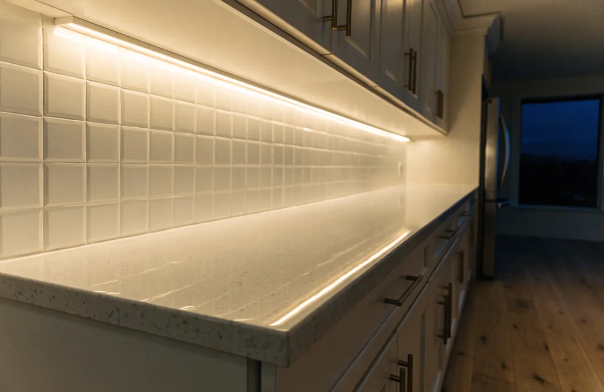 The counter run of that Davie kitchen seen along its length after dark, the under-cabinet light running the whole length of the stone with the cabinet doors above lit only by the spill