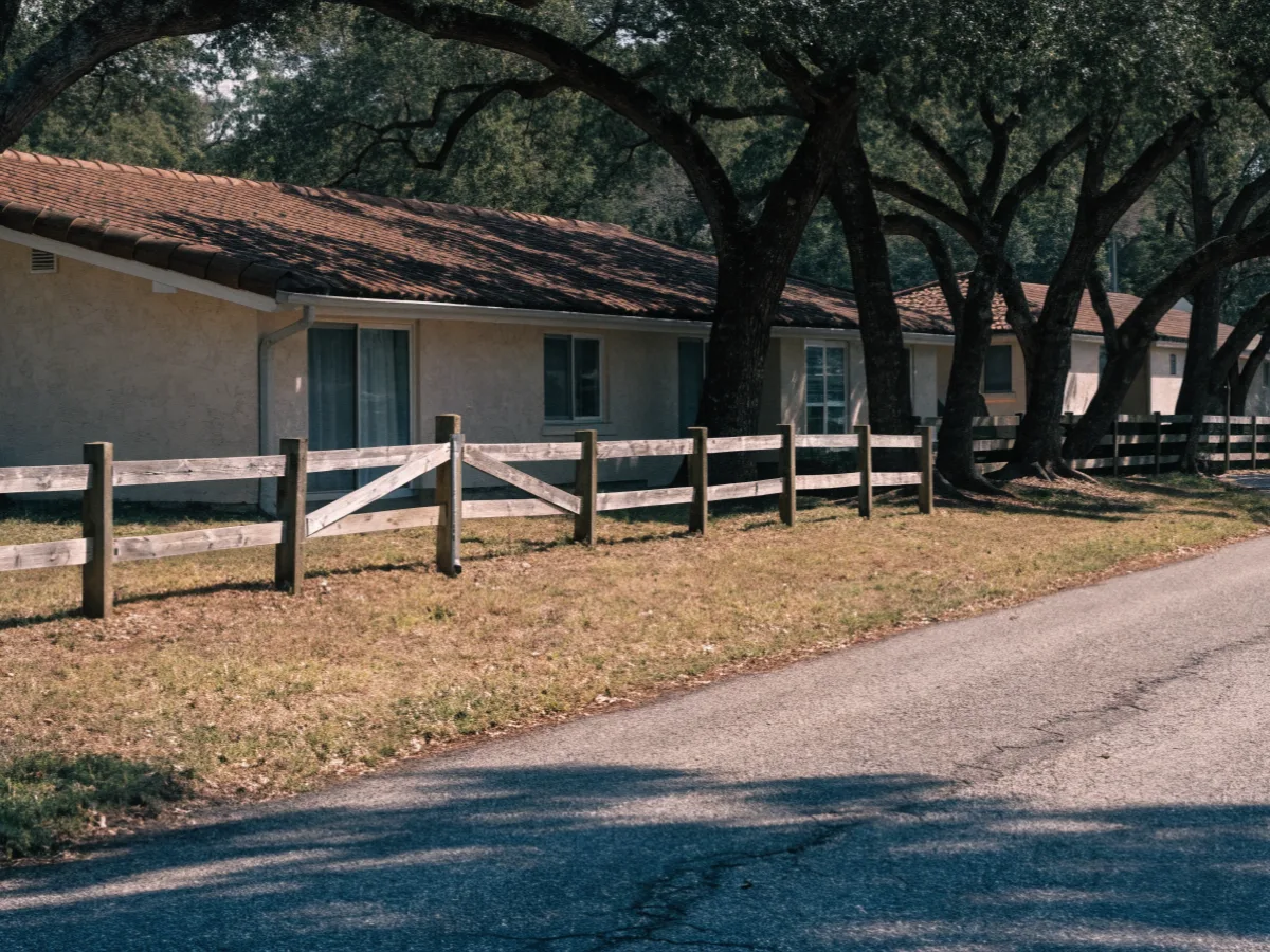 A four foot split rail fence on the front line of a 1970s single story house in Davie, mature oaks shading the two lane street behind it