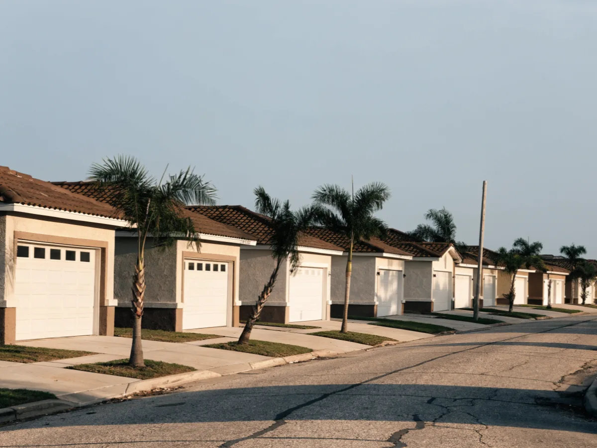 A 1990s subdivision street in west Davie, single story houses with two car garages, concrete driveways and young palms along the swale