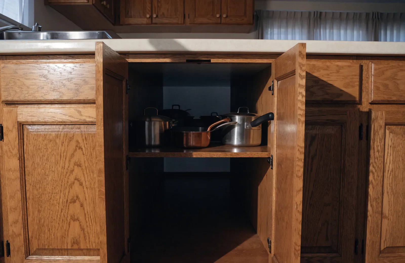 A deep base cabinet in a 1980s Davie kitchen with one fixed shelf, the front row of pots lit and the back of the shelf lost in shadow behind them