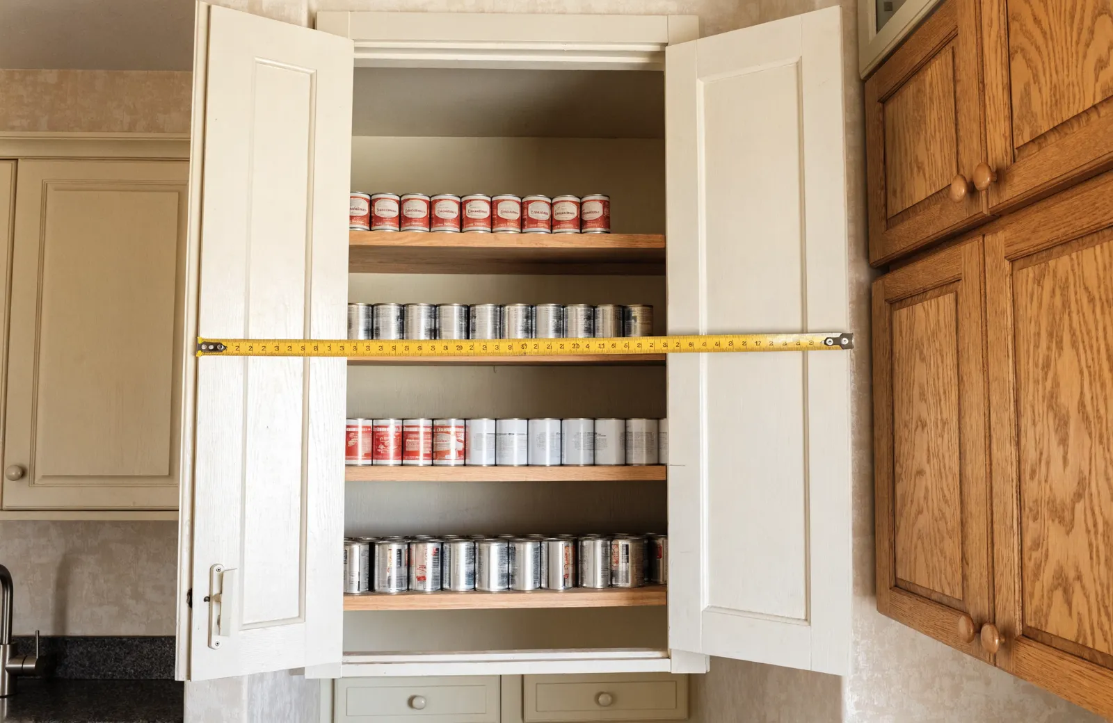 The pantry closet of a 1987 Davie kitchen measured with a tape across the opening, the fixed shelves inside stacked with cans one row deep